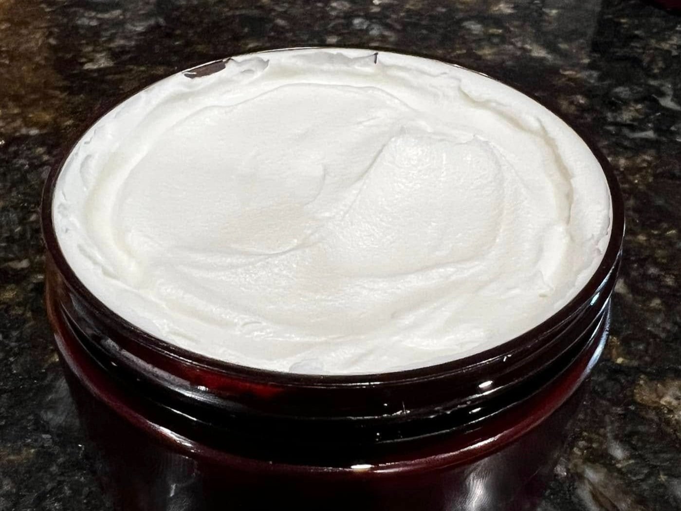 480697070_122108924366741856_7279774347555266242_n A close-up view of a luxurious, creamy skincare moisturizer in a dark brown jar, featuring a smooth, whipped texture on top, placed on a dark granite surface.