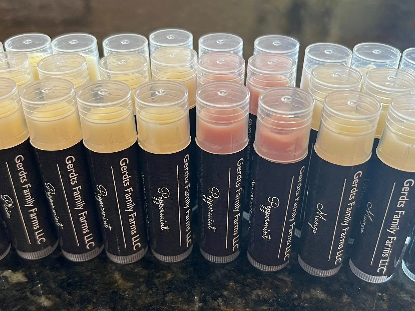 Gerdts+Family+Farms+LLC+-+494698163_122123222060741856_2014898171784469176_n Array of lip balm tubes from Gerdts Family Farms LLC, featuring flavors like Peppermint and Mango, displayed on a dark countertop. Ideal for natural skincare enthusiasts seeking moisturizing solutions.