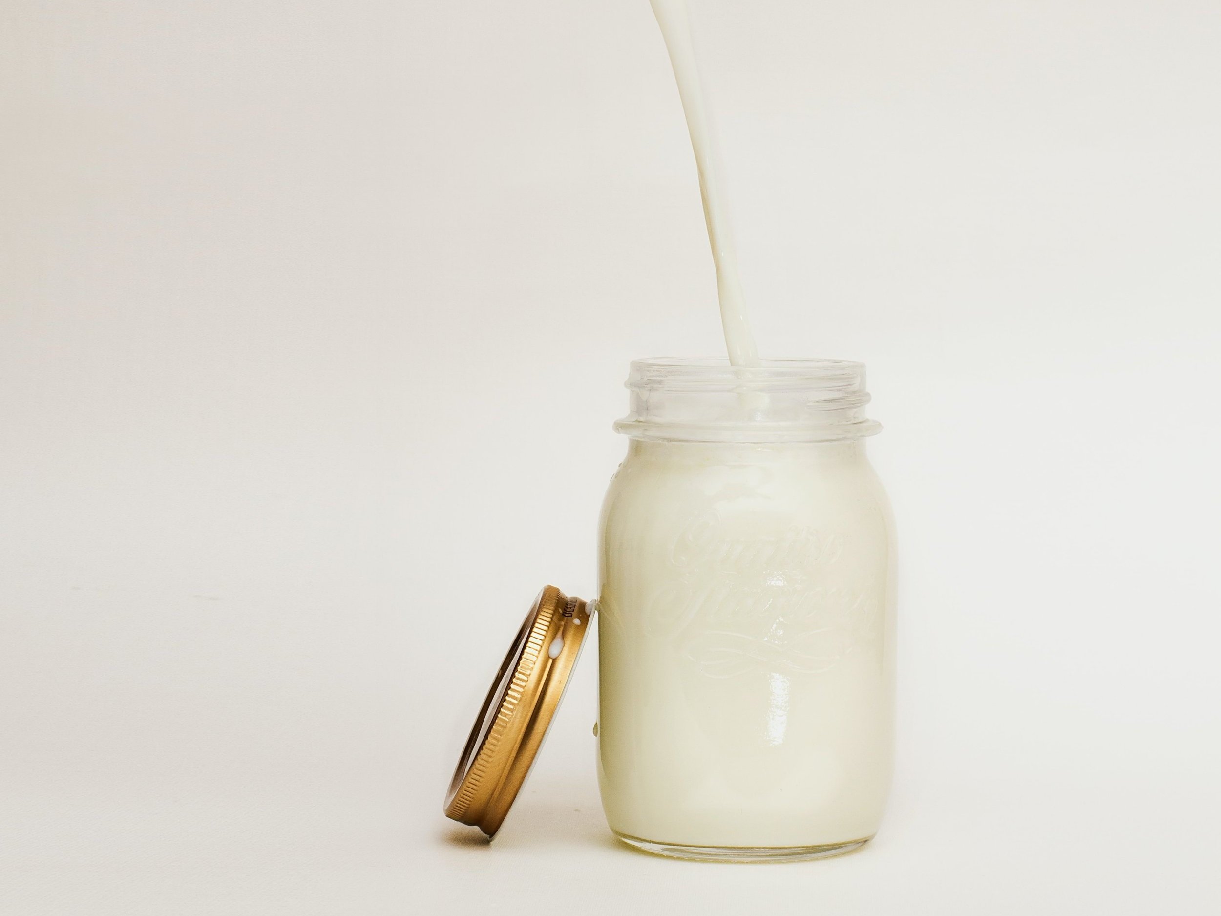 milk_jar (2) Creamy milk being poured into a clear glass jar with a gold lid, set against a soft white background, highlighting freshness and quality.